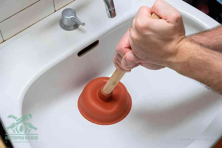 Twee handen gebruiken een rode rubberen plopper in een witte wastafel om de verstopte afvoer los te pompen in Rijswijk