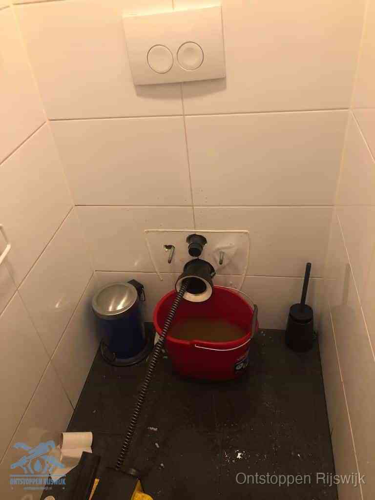 Een ontstopte wand-wc met verwijderde toiletpot waar een ontstoppingsveer in de wanduitgang verdwijnt, het achterhalen waar de verstopping zit in Rijswijk
