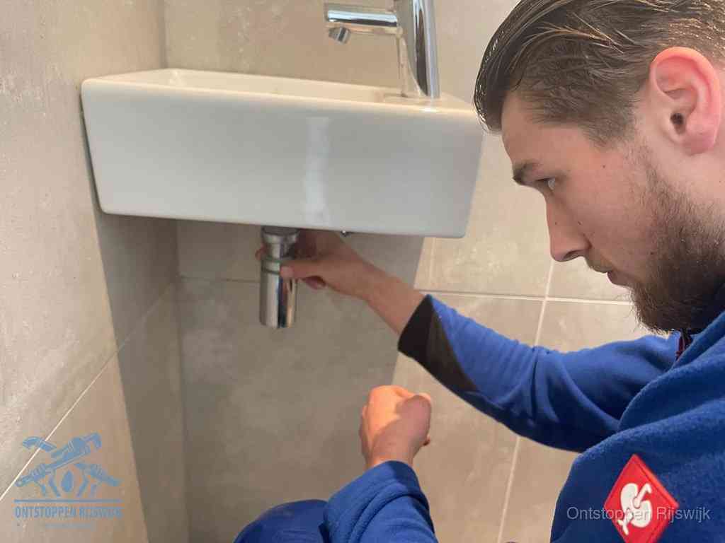 Een monteur draait met de hand de zwanenhalssifon onder een fonteinwasbak in de toiletruimte los om de bron van rioollucht in Rijswijk te vinden