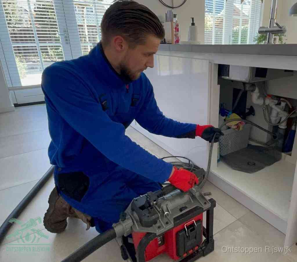 Een monteur met rode handschoenen knielt in een keuken en voert een elektrische ontstoppingsveer in tijdens het professioneel afvoer ontstoppen in Rijswijk