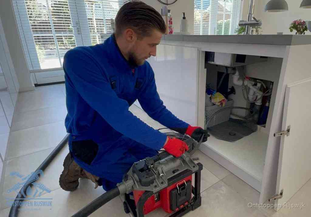 Een monteur voert een professionele elektrische ontstoppingsmachine in de afvoer onder de spoelbak tijdens het gootsteen ontstoppen in Rijswijk